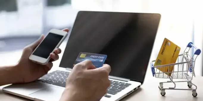 Hands holding a phone and a credit card at a laptop, beside a miniature shopping trolley