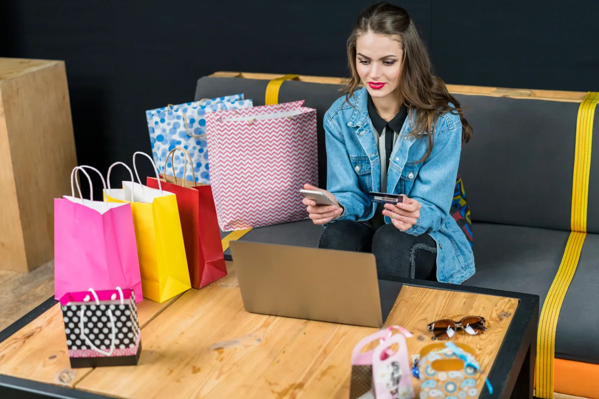 A woman on a sofa surrounded by shopping bags, paying for an order online with her card and laptop