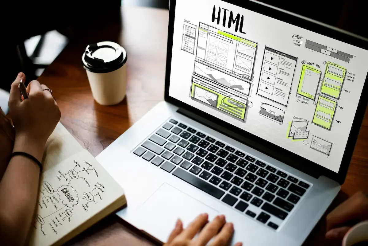A laptop showing hand-drawn HTML wireframes beside a brand-planning notebook